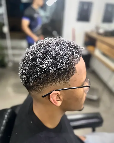 Cliente com luzes platinadas (nevou) na cadeira, com o barbeiro trabalhando ao fundo
